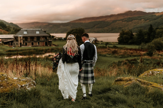 Bride wearing leather jacket and groom wearing kilt walk through grass